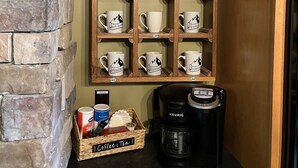 Um espaço para café com parede de pedra, prateleira de madeira com canecas, cafeteira e um cesto com café e chá.