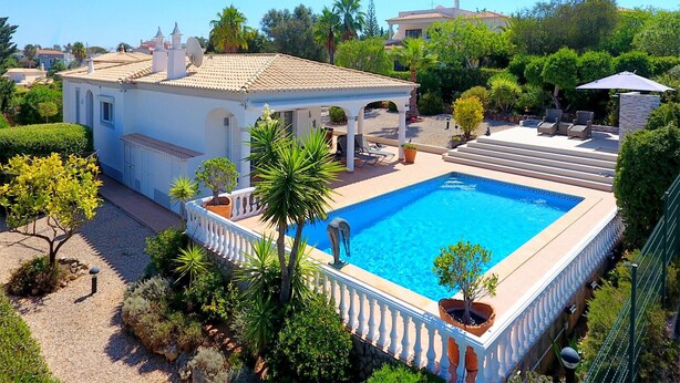 Uma grande piscina com balaustrada branca, rodeada por vegetação exuberante e um edifício branco com telhado ladrilhado.