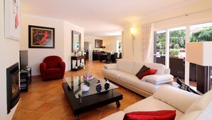 Uma sala de estar com sofá, poltrona e mesa de centro. Ao fundo, há uma área de jantar com uma mesa e cadeiras.
