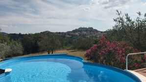Piscina azul y transparente con borde curvo, rodeada de vegetación y vistas a un pueblo lejano.