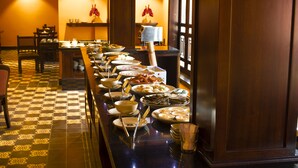A buffet setup with various dishes and utensils on a long table, surrounded by chairs and tables in a warmly lit room with patterned flooring. - Spatel d'Annam Boutique Spa & Hotel (Hue)