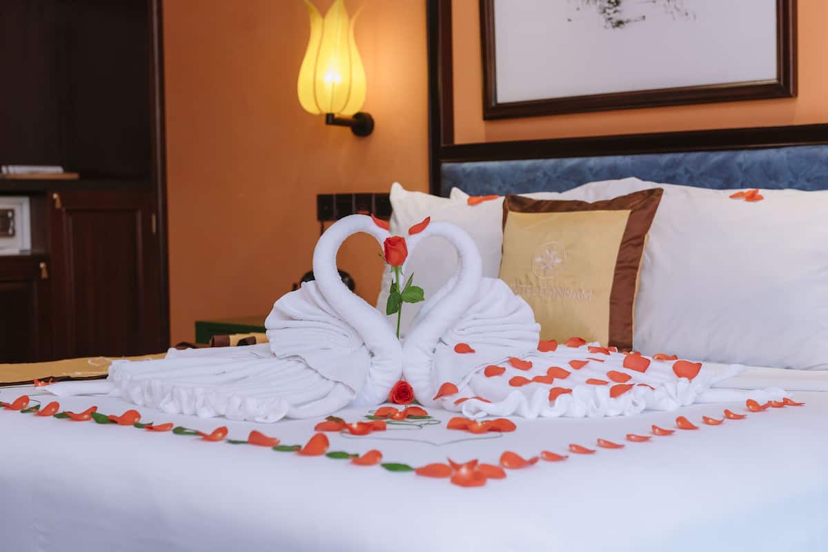 Una cama impecable con una toalla dispuesta en forma de corazón y una rosa, rodeada de pétalos de rosa.