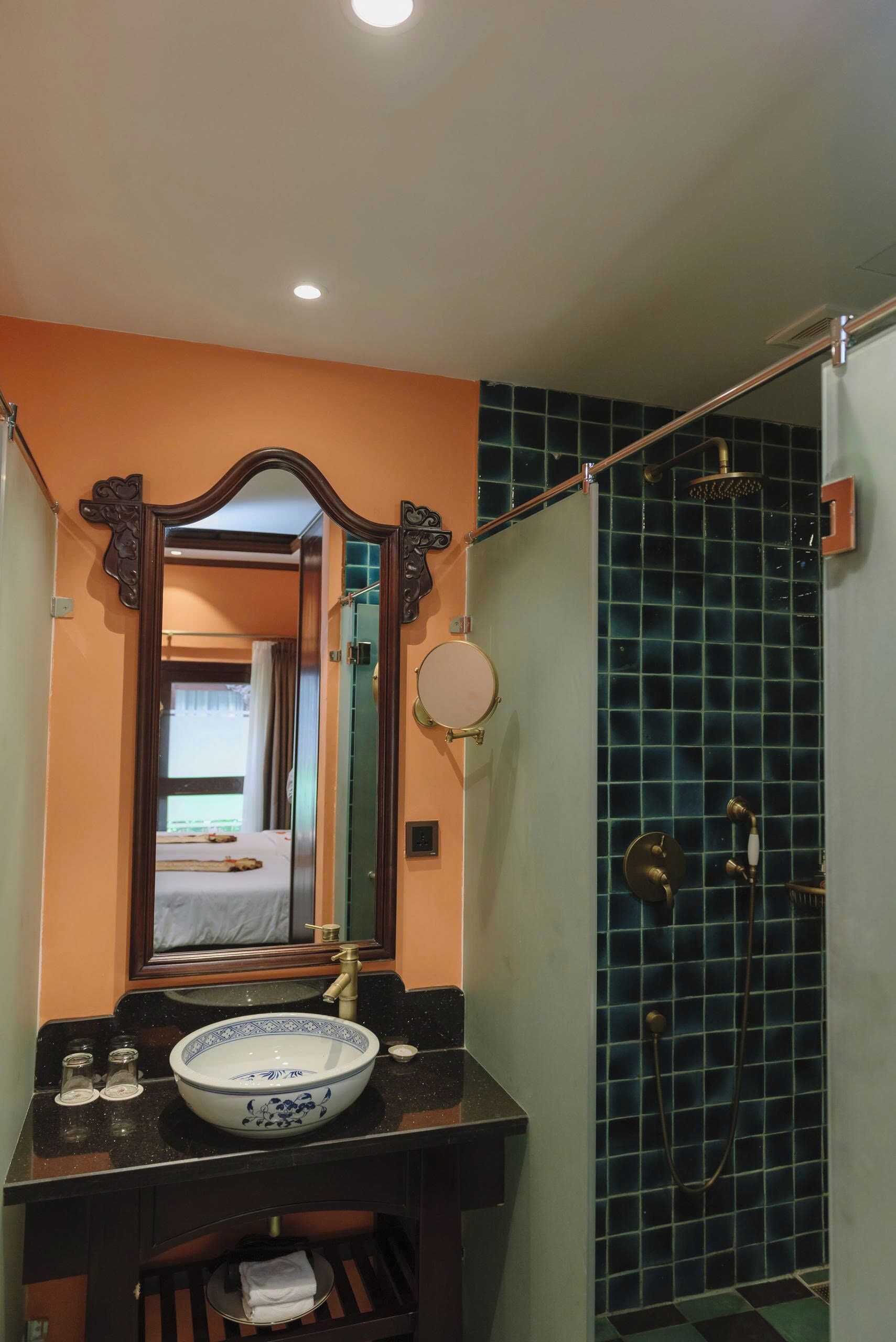 A bathroom with a large mirror, a sink with a decorative bowl, a shower with blue tiles, and a door leading to a bedroom.