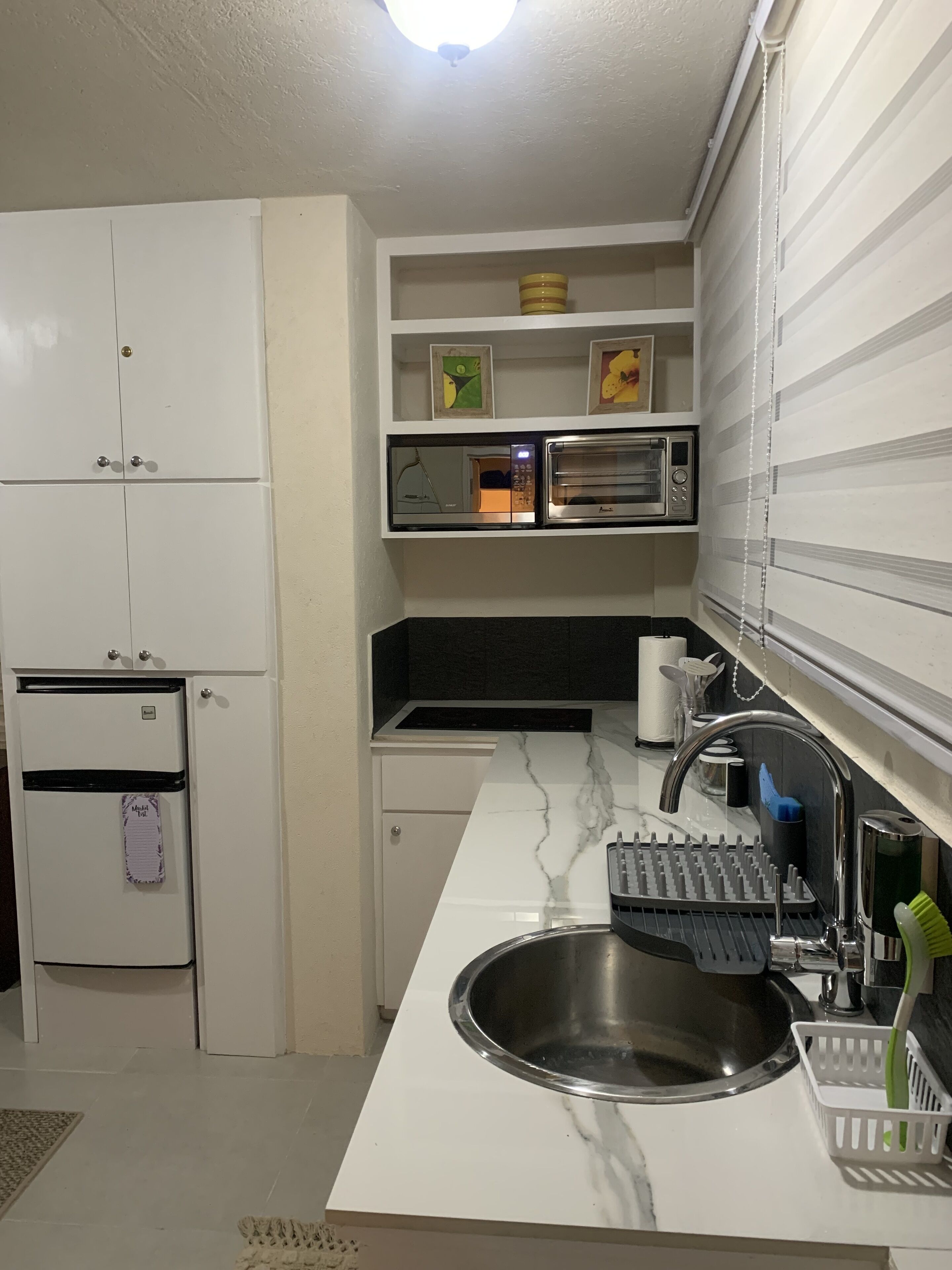 A compact kitchen with white cabinets, a built-in microwave, a stainless steel sink, and a small refrigerator.