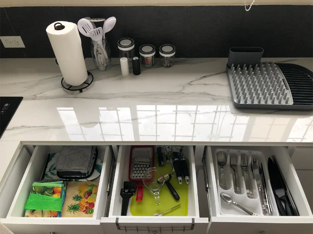 A kitchen counter with a paper towel holder, spatulas, jars, and a grater. Below, a drawer contains a wallet, a green cloth, a glass, and utensils.