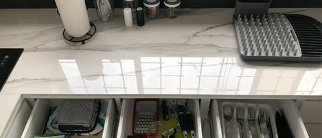 A kitchen counter with a paper towel holder, spatulas, jars, and a grater. Below, a drawer contains a wallet, a green cloth, a glass, and utensils.