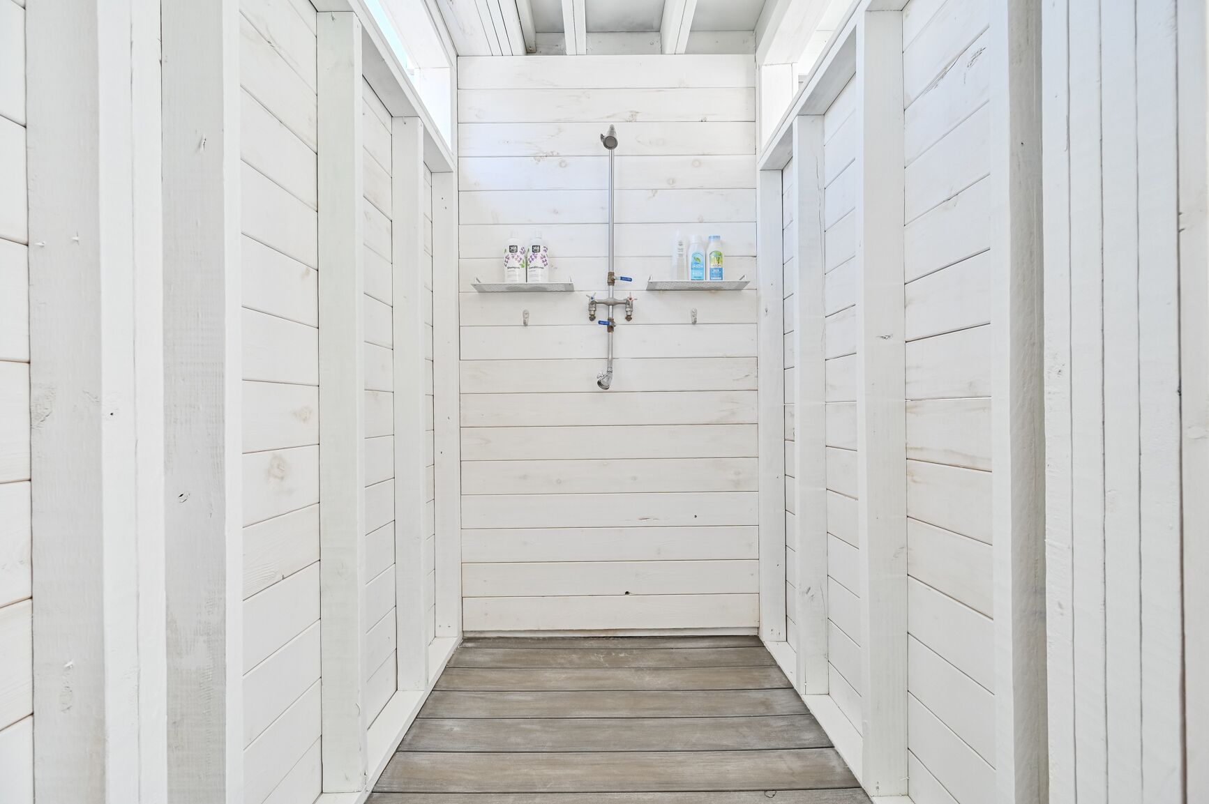 A narrow hallway with white wooden walls and a wooden floor, leading to a shower area with a showerhead and two shelves holding toiletries.