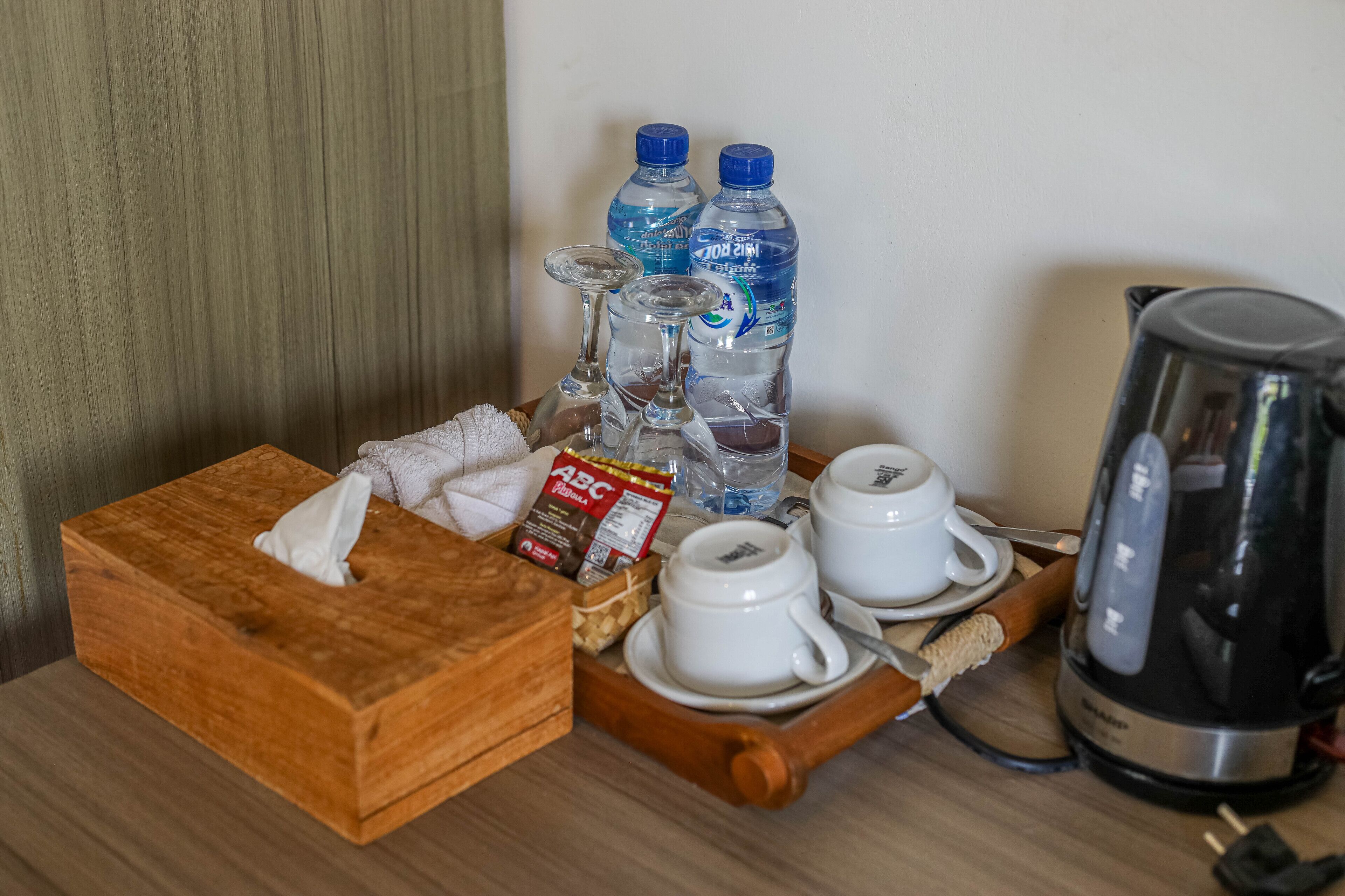 A wooden tray with a tissue box, two cups, a kettle, and two bottles of water on a wooden table.
