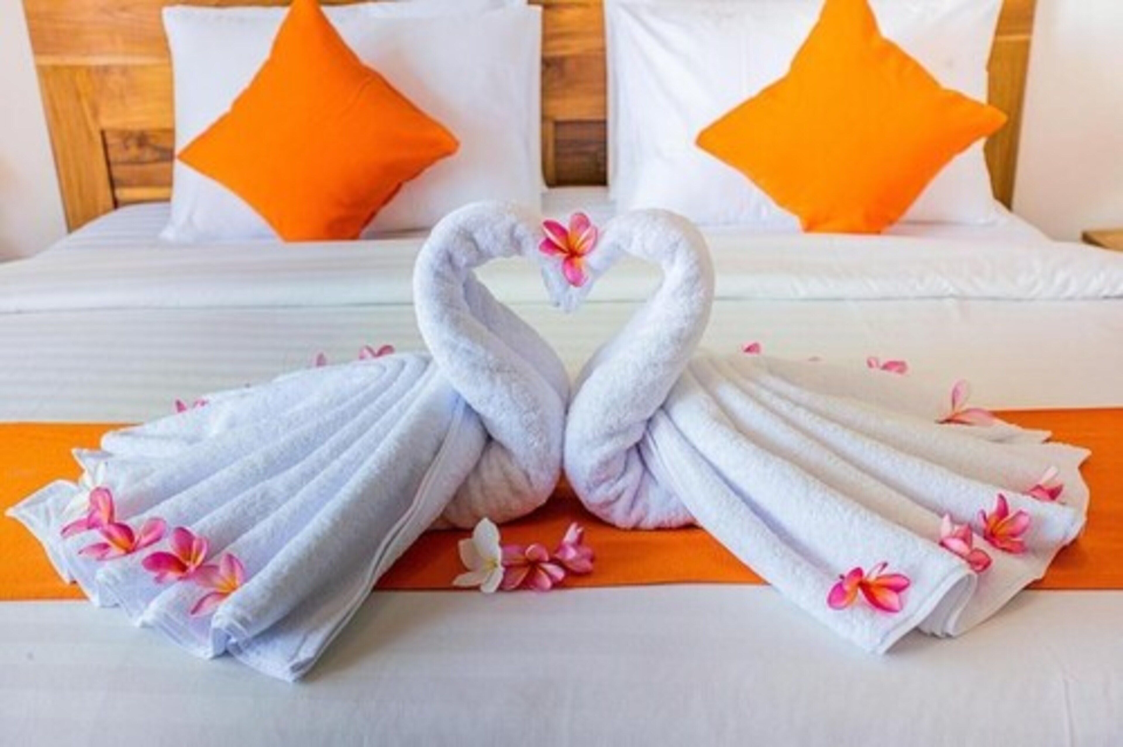 A bed with white bedding and orange pillows, a heart-shaped towel arrangement, and a floral decoration.