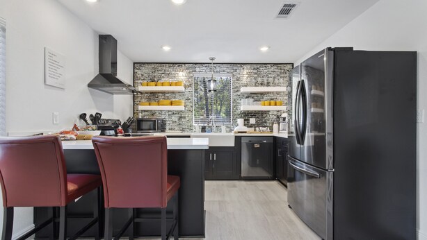 A modern kitchen with a black refrigerator, two red bar stools, a central island, and a backsplash with a cityscape view. - Vibrant & Enchanting! Hot Tub! Walk to the Beach! (Pass Christian)