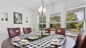 A dining area with a round table set for tea service, surrounded by leather chairs, large windows with blinds, and a chandelier. - Vibrant & Enchanting! Hot Tub! Walk to the Beach! (Pass Christian)