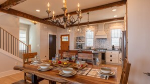 A dining area with a wooden table set for six, a chandelier, and a kitchen in the background. - Blue Ridge Bliss - BRMC amenities, views, firepit, indoor and outdoor gas fireplace (Boone)