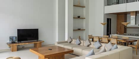 A modern living room with a large sofa, wooden coffee table, and wicker ottomans. There is a flat-screen TV on a wooden stand, and a dining area with a wooden table and chairs.