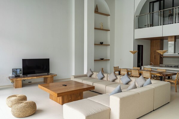 A modern living room with a large sofa, wooden coffee table, and wicker ottomans. There is a flat-screen TV on a wooden stand, and a dining area with a wooden table and chairs. - Lavender Rooftop 3BR Villa with a Heated Private Pool (Cimenyan)
