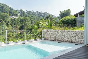 A swimming pool with a stone wall and metal fence, surrounded by lush greenery and a view of a hillside with houses.