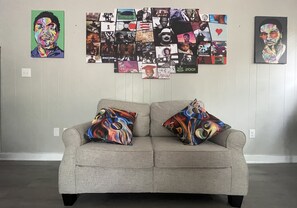 Ein Wohnzimmer mit grauem Sofa und bunten Kissen, einer Bildercollage an der Wand und einer schlichten weißen Wand.