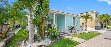 Una casa residencial con porche, rodeada de plantas tropicales y un césped bien cuidado.