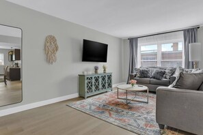 A modern living room with a grey sofa, a coffee table, a flat-screen TV, and a decorative macrame wall hanging.