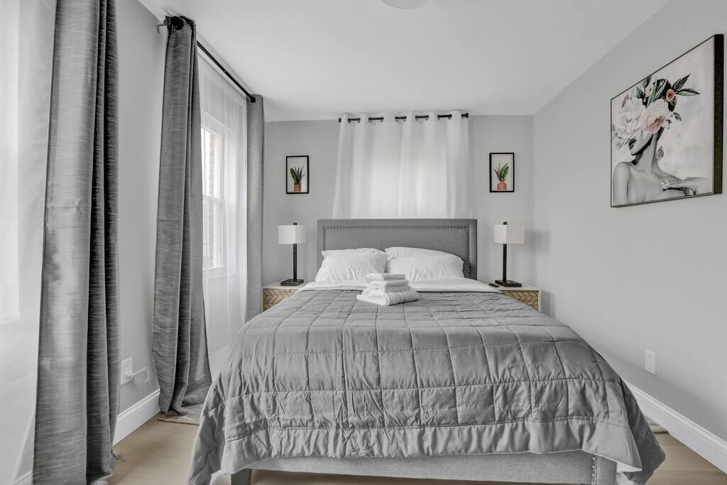 A neatly arranged bedroom with a large bed, two bedside tables with lamps, a window with sheer curtains, and a framed artwork on the wall.
