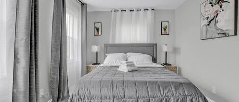 A neatly arranged bedroom with a large bed, two bedside tables with lamps, a window with sheer curtains, and a framed artwork on the wall.