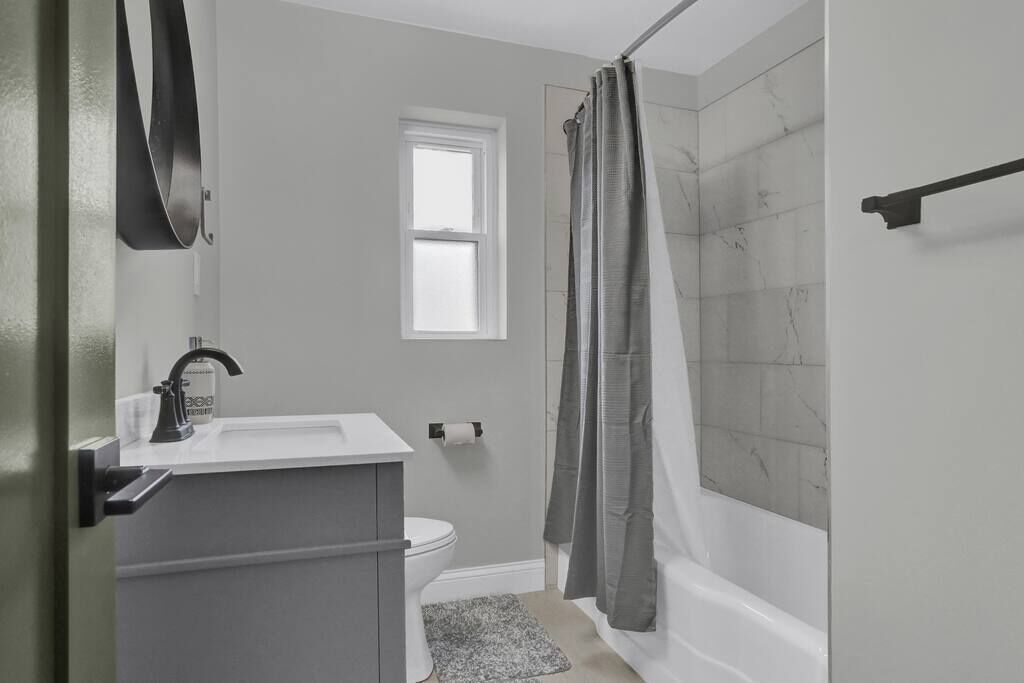 Ein Badezimmer mit weißem Waschbecken, Toilette, Dusche mit grauer Duschvorhang und einem Fenster.