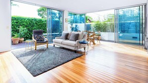 A modern living room with a sofa, chairs, a coffee table, and a large glass door leading to a balcony with a view of greenery. - The Garden Retreat (Bondi Junction)