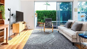 A modern living room with a flat-screen TV, a sofa, a coffee table, and a large sliding glass door leading to a balcony with plants. - The Garden Retreat (Bondi Junction)