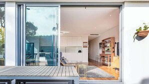 A modern house with a sliding glass door leading to a patio with a wooden table and chairs. Inside, there is a kitchen with white cabinets and a dining area with a sofa and a coffee table. - The Garden Retreat (Bondi Junction)