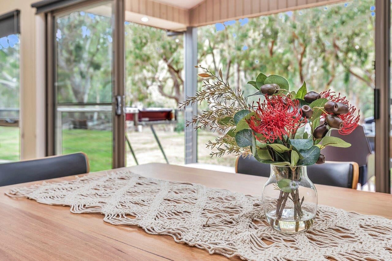 A table with a lace tablecloth, a vase with flowers, and a view of a garden through large windows.