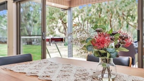 A table with a lace tablecloth, a vase with flowers, and a view of a garden through large windows. - Luxury Waterfront Retreat on Murray River (Moama)