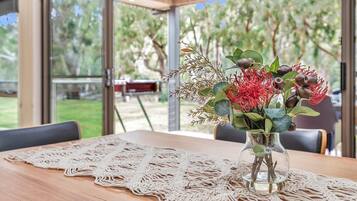 A table with a lace tablecloth, a vase with flowers, and a view of a garden through large windows.