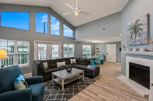 A spacious living room with a large sofa, a coffee table, and a fireplace. There are large windows and a ceiling fan.