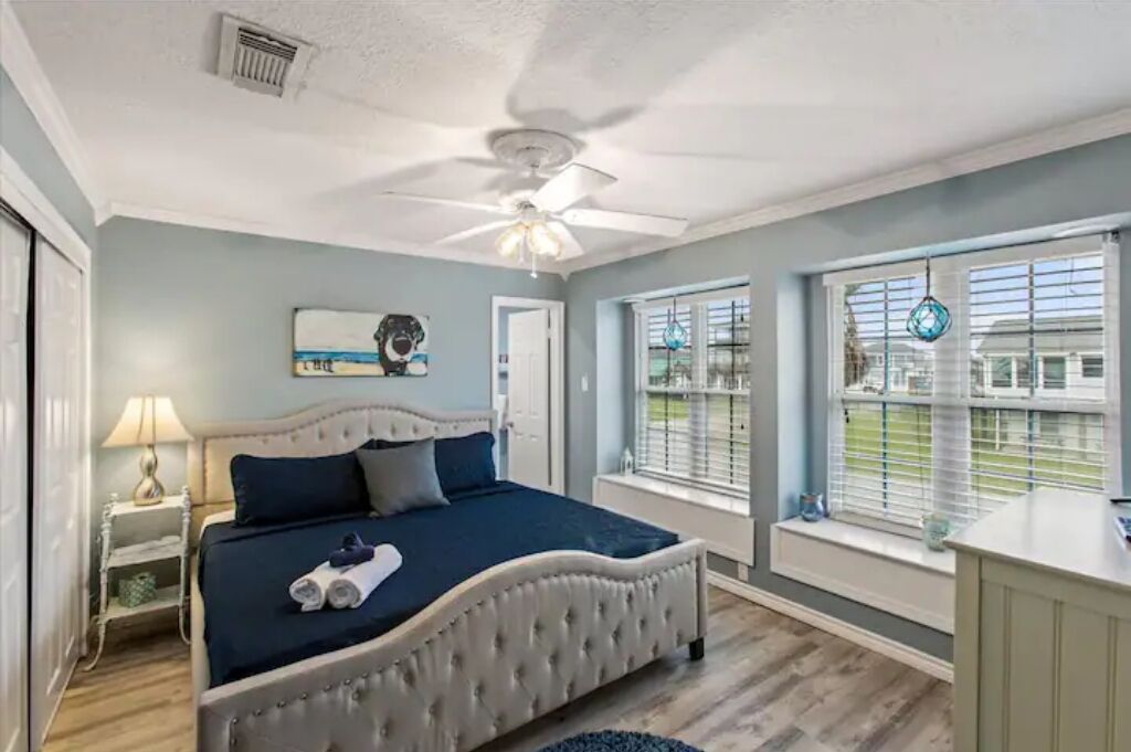 A bedroom with a bed, a ceiling fan, a window with blinds, and a nightstand with a lamp.