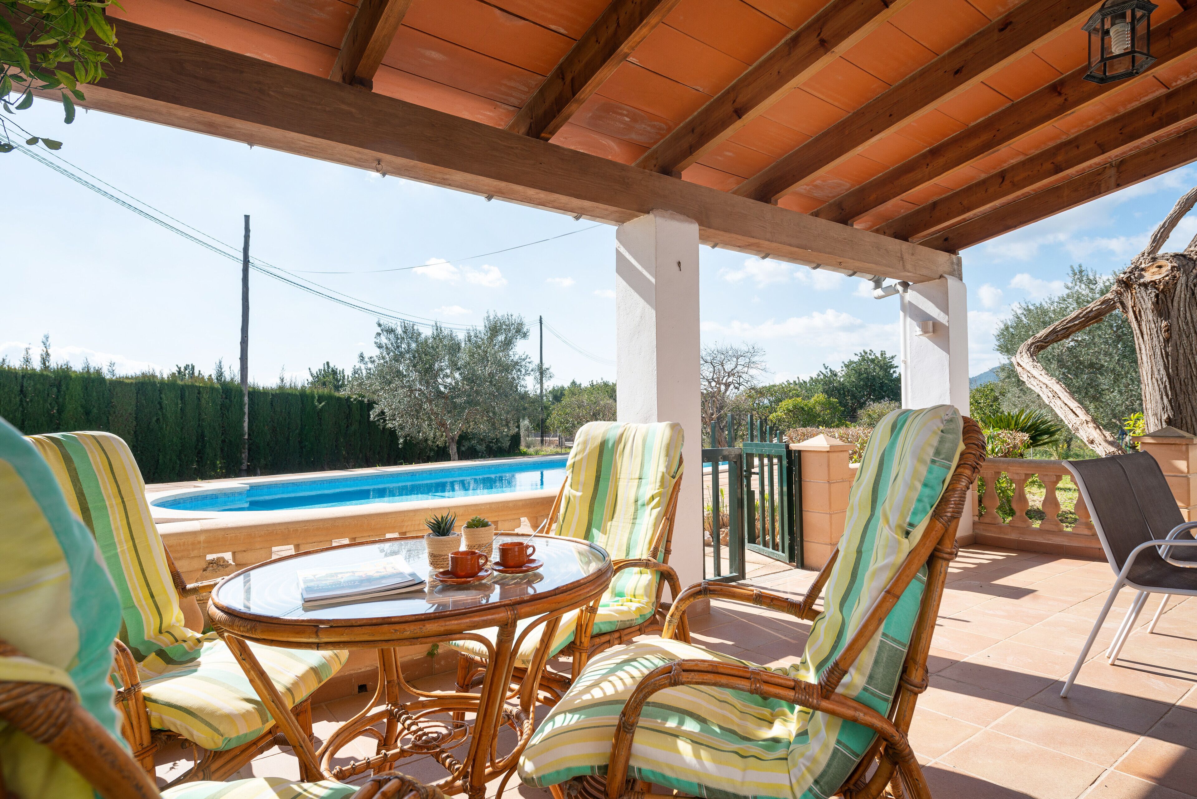 Un área de estar al aire libre techada con muebles de mimbre, una mesa de vidrio y vista a una piscina.