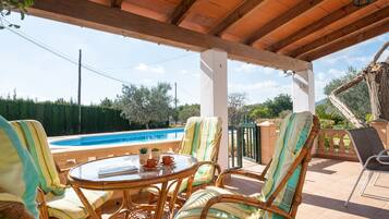 Un área de estar al aire libre techada con muebles de mimbre, una mesa de vidrio y vista a una piscina.