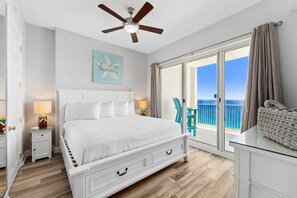 Una camera da letto con un letto grande, un ventilatore a soffitto, vista sull'oceano tramite una porta scorrevole in vetro e un tavolino con una borsa sopra.
