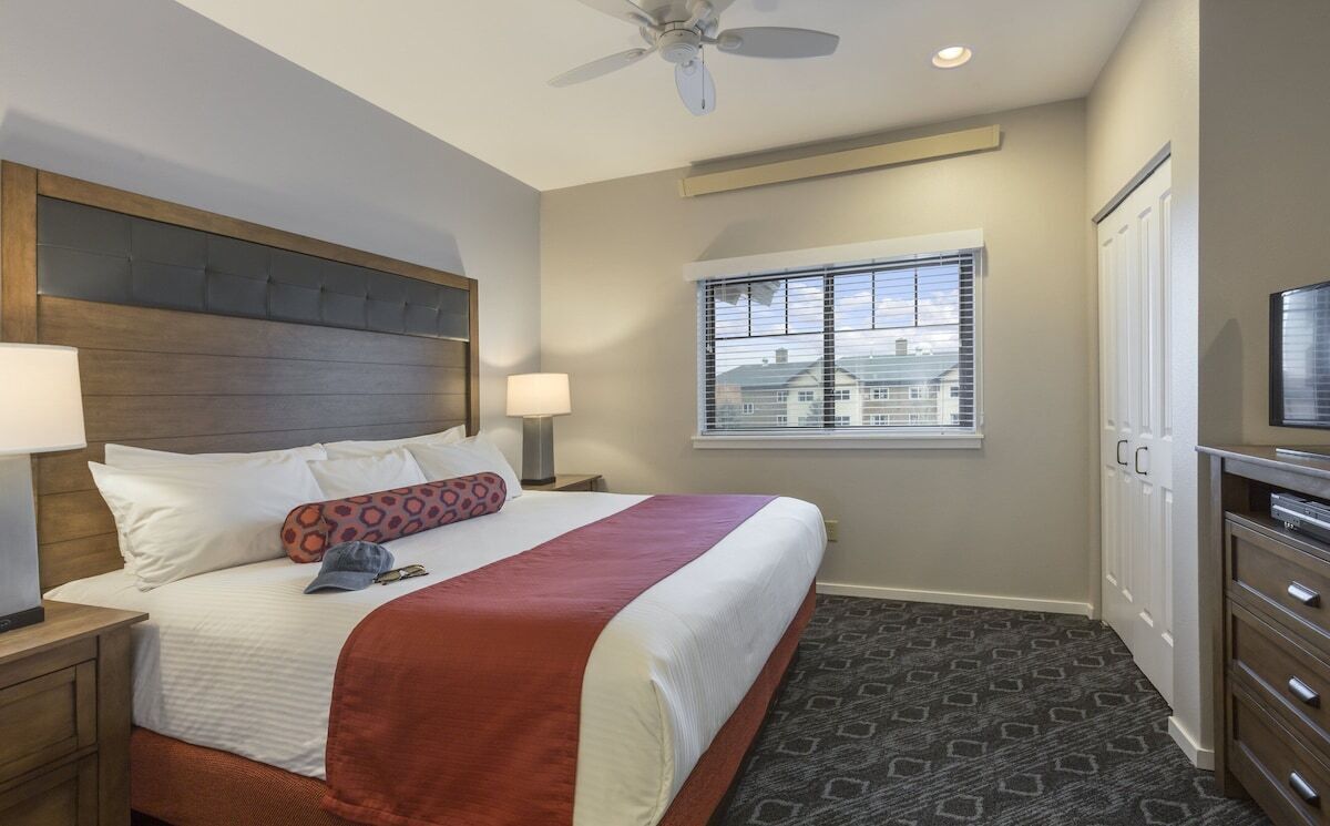A hotel room with a large bed, a ceiling fan, a window with blinds, a TV, and a wardrobe.