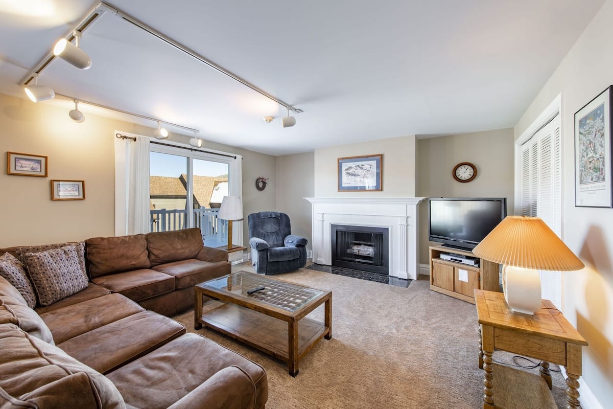 A living room with a brown leather sofa, a wooden coffee table, a fireplace, a television, and a lamp on a side table.