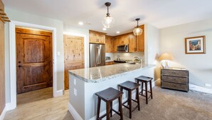A modern kitchen with wooden cabinets, a stainless steel refrigerator, a microwave, and a granite countertop. There are three bar stools and a small island with a sink. - Resort Plaza Condominiums 5030 - 5064 (Park City)