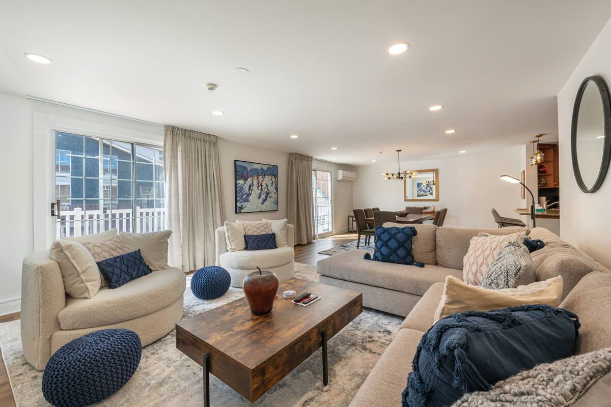 A spacious living room with a large window, a sofa set, a coffee table, and a dining area in the background.
