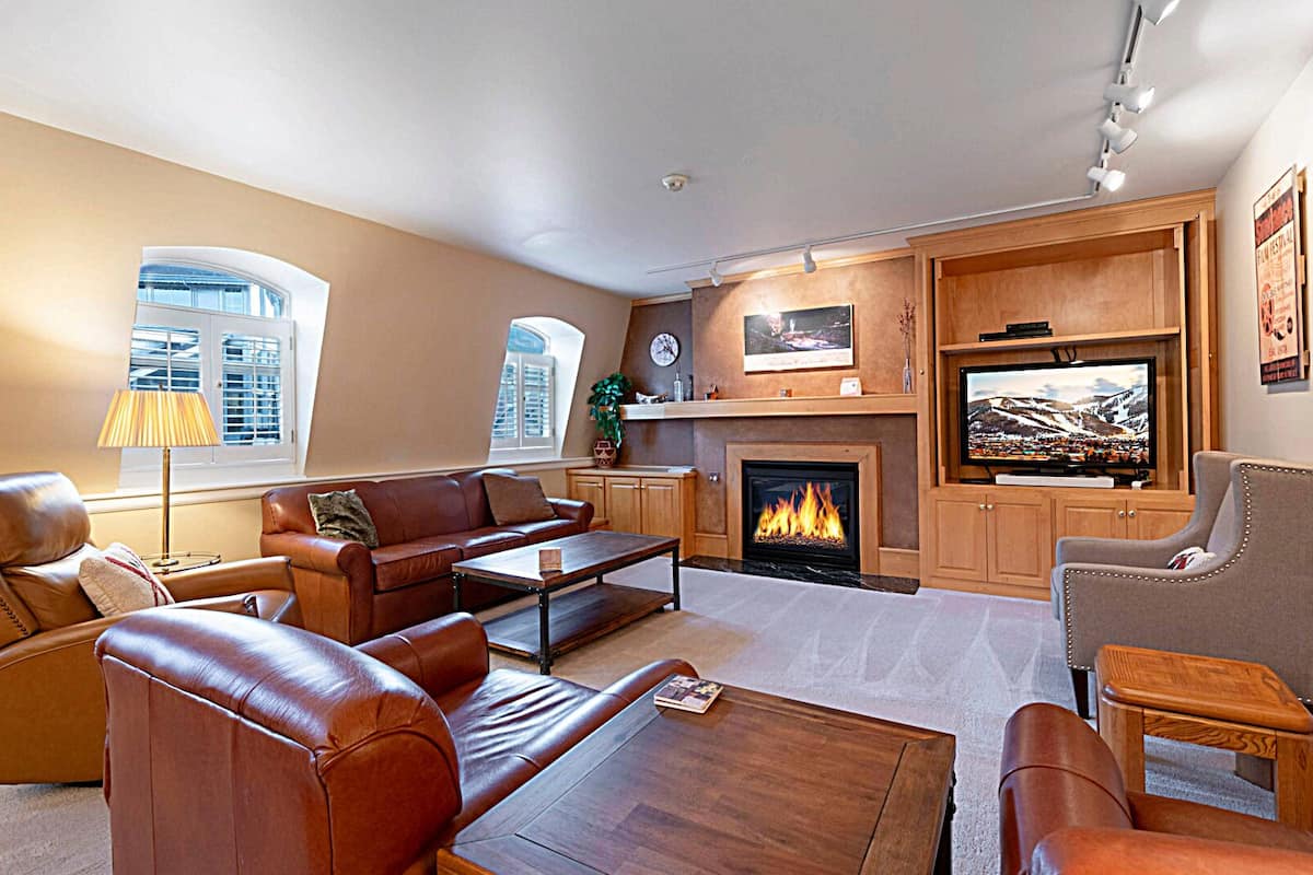 A cozy living room with leather sofas, a fireplace, a flat-screen TV, and a large window with blinds.