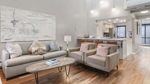 A modern living room with a sofa, armchairs, and a coffee table. There is a kitchen area with a dining table and chairs visible in the background. - Park Avenue Upgraded 2 Bedroom Condominiums (Park City)