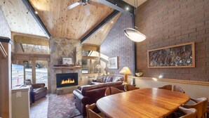 A cozy living room with a fireplace, leather sofas, a dining table, and a large window overlooking a snowy landscape. - Park Avenue Upgraded 2 Bedroom Condominiums (Park City)
