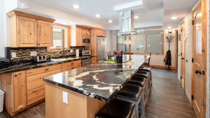 A modern kitchen with wooden cabinets, a large island with bar stools, stainless steel appliances, and a large window providing natural light. - Park Avenue Upgraded 2 Bedroom Condominiums (Park City)