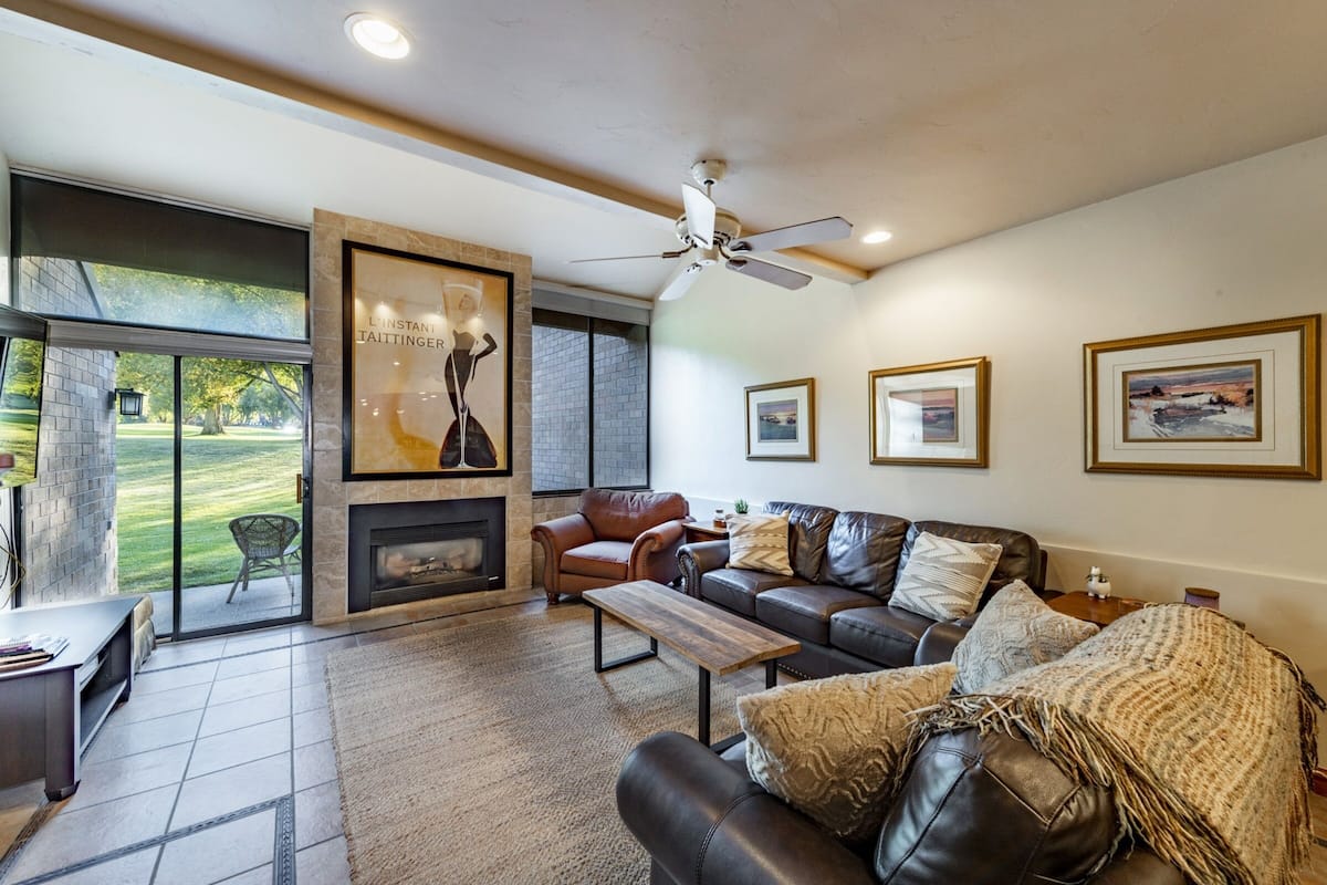 A spacious living room with a fireplace, leather sofas, a coffee table, and large windows offering a view of the outdoors.