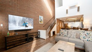 A modern living room with a large flat-screen TV, wooden flooring, a staircase, a sofa, and a coffee table. - Park Avenue 3 Bedroom Condominiums (Park City)