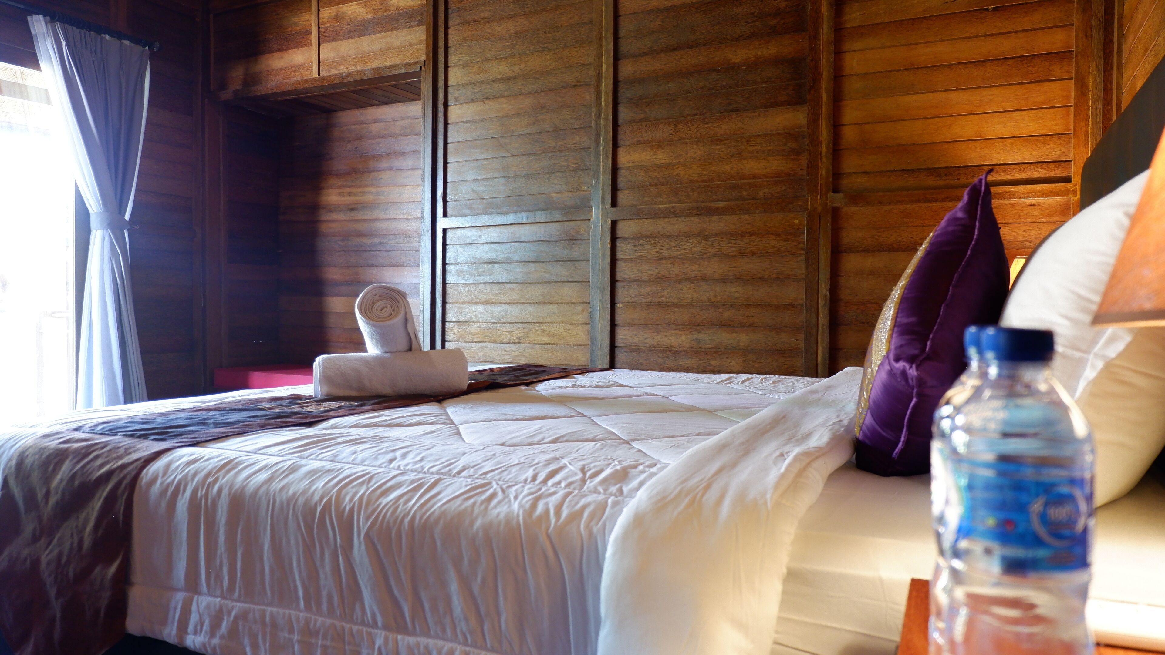 A neatly made bed with a rolled towel, a water bottle, and a purple pillow.