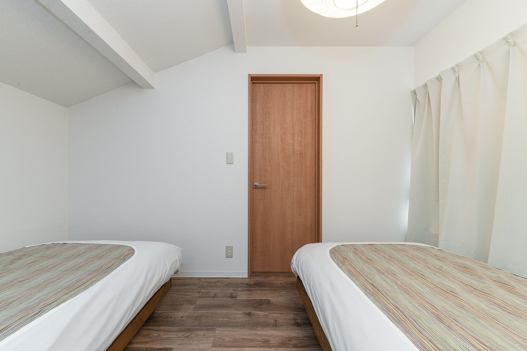 A room with two single beds, a wooden door, and a light switch on the wall.