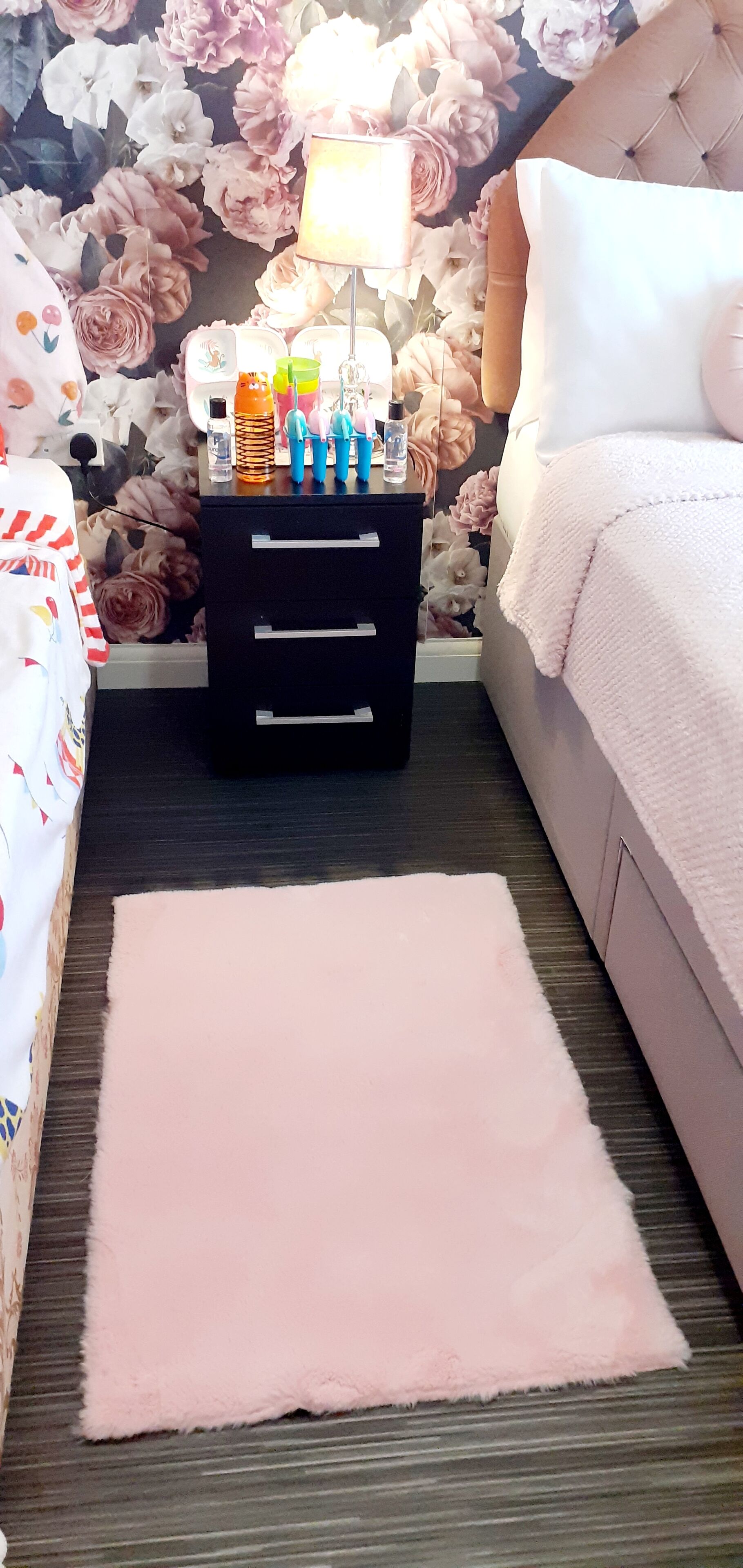 A bedroom with a floral wallpaper, a bedside table with toiletries, a fluffy pink rug, and a bed with a white blanket.