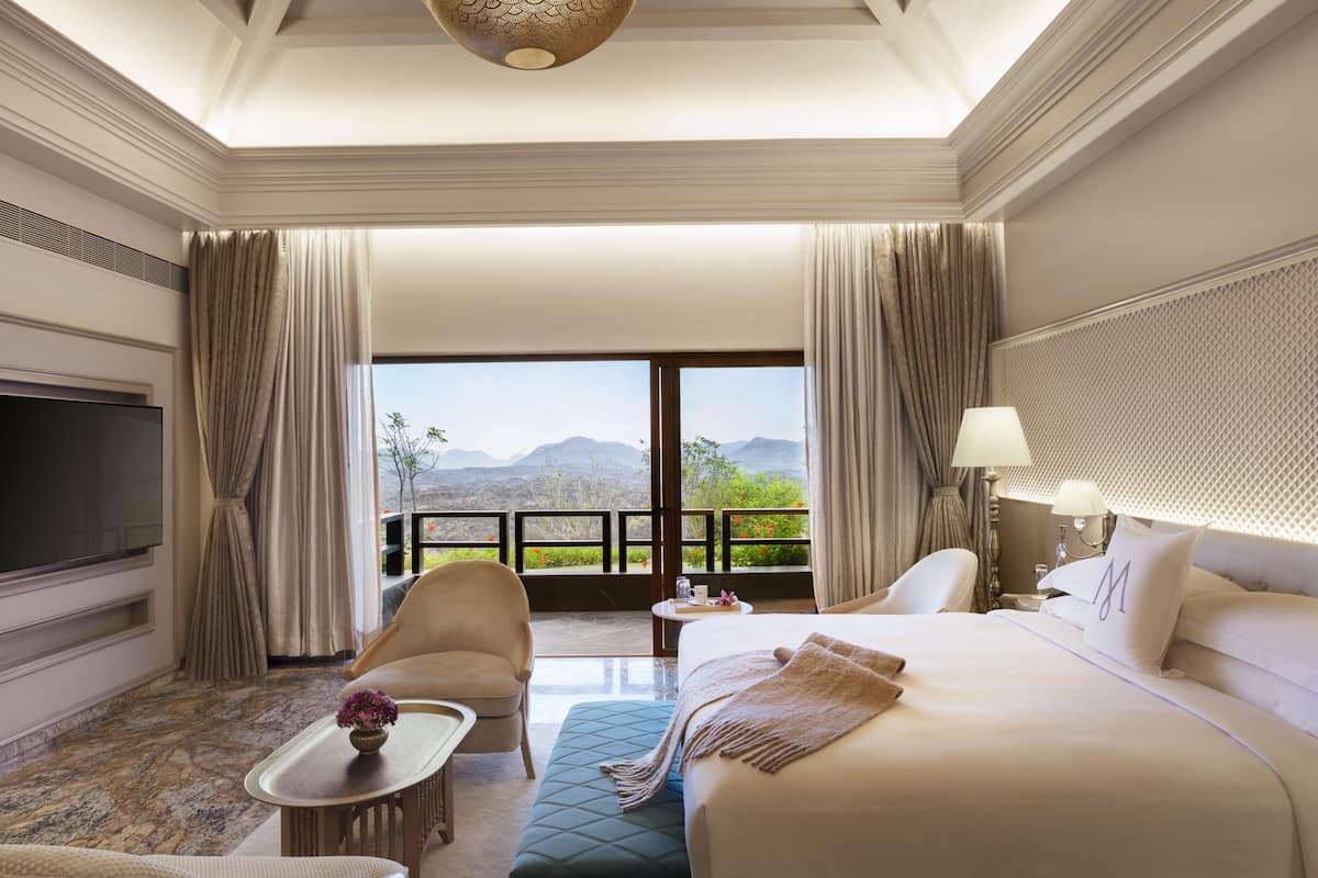 A spacious hotel room with a large bed, a television, a seating area with a table, and a balcony with a mountain view.
